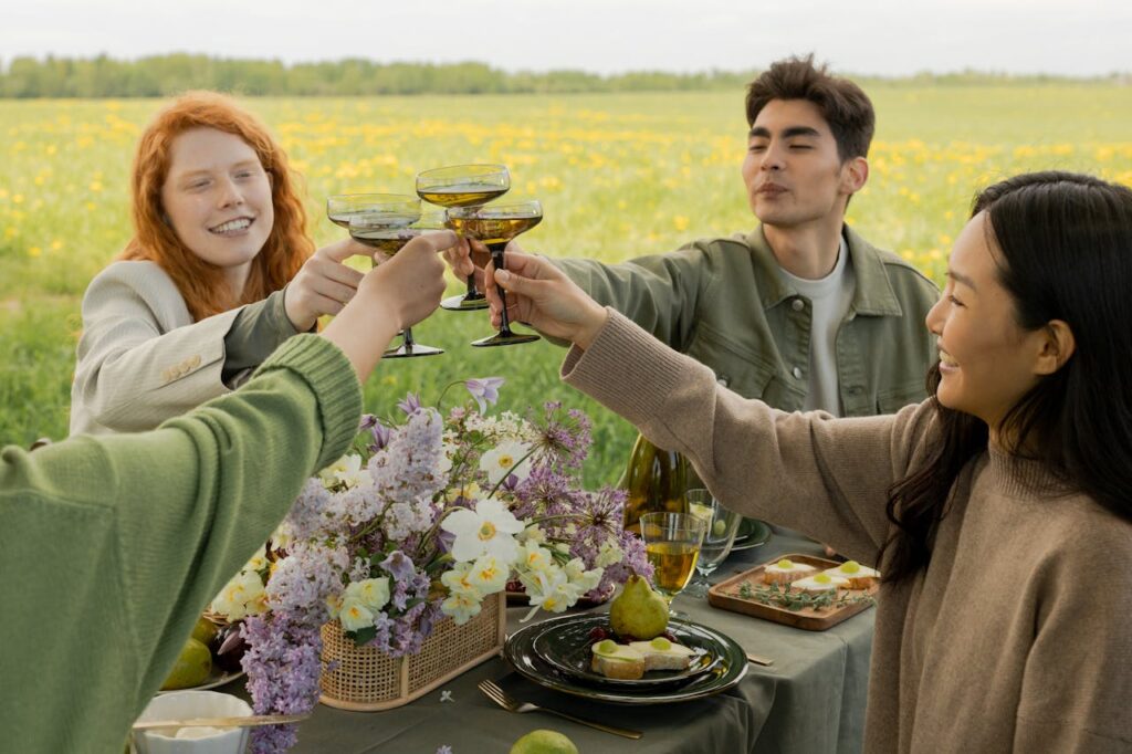 pexels-photo-9577524 Friends enjoying a cheerful outdoor picnic, raising glasses in a meadow setting with flowers and food.